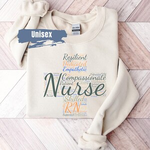 May include: A beige unisex sweatshirt with a word cloud design featuring the word "Nurse" in large, dark green letters. The word cloud includes words like "resilient", "dedicated", "compassionate", "patient", "caring", "skilled", and "RN".