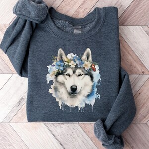 Op de afbeelding: Een donkergrijze sweatshirt met een aquarel illustratie van een husky hond met een bloemenkrans. Het gezicht van de hond is gedetailleerd met grijstinten, zwart en wit, tegen een blauwe aquarel achtergrond.