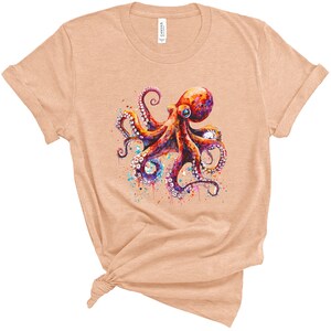 Puede incluir: Una camiseta de color melocot&oacute;n claro con una ilustraci&oacute;n de acuarela de un pulpo con tent&aacute;culos coloridos y una salpicadura de pintura alrededor.