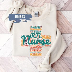 May include: A beige unisex sweatshirt with a teal and orange word cloud design that says "RN Nurse" with words like "reliable", "compassionate", "patient", "skilled", "dedicated", and "resilient".
