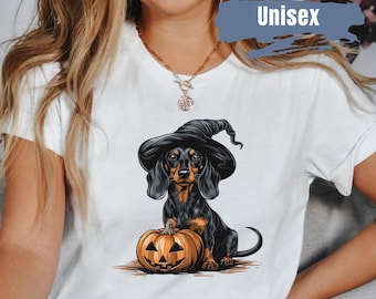 Op de afbeelding: Wit unisex T-shirt met een Halloween-ontwerp. Het toont een zwart-bruine teckel met een heksenhoed, naast een uitgesneden pompoen. Het woord "Unisex" staat rechtsboven op het shirt.
