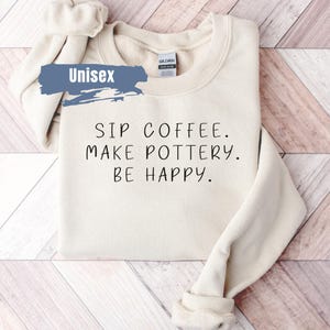 May include: A cream-colored unisex sweatshirt with the text "SIP COFFEE. MAKE POTTERY. BE HAPPY." printed in black.