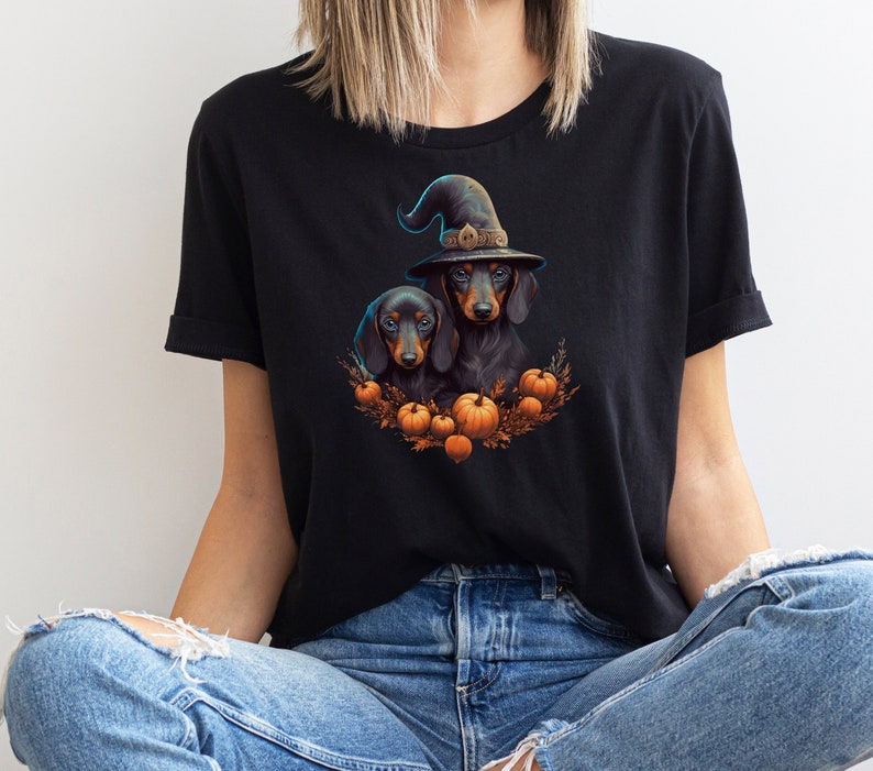 Może przedstawiać: Czarny t-shirt z ilustracją na Halloween. Grafika przedstawia dwa jamniki, z kt&oacute;rych jeden ma na sobie kapelusz czarownicy, otoczone dyniami i jesiennymi liśćmi. Projekt jest kolorowy i szczeg&oacute;łowy, idealny na straszny sezon.