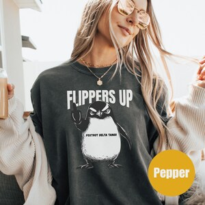 May include: A dark gray sweatshirt with a graphic of a penguin with its flippers up. The penguin is wearing a hat that says "Fox Trot Delta Tango". The text "Flippers Up" is printed above the penguin.