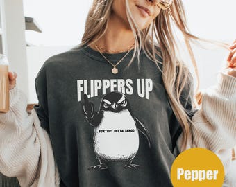 Puede incluir: Una sudadera gris oscuro con un gráfico de un pingüino con las aletas levantadas. El pingüino lleva un sombrero que dice "Fox Trot Delta Tango". El texto "Flippers Up" está impreso sobre el pingüino.