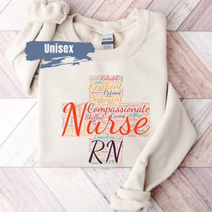 May include: A beige unisex sweatshirt with a word cloud design featuring the word "Nurse" in large, orange letters. The word cloud includes words like "reliable", "resilient", "patient", "dedicated", "compassionate", "skilled", "caring", and "selfless".