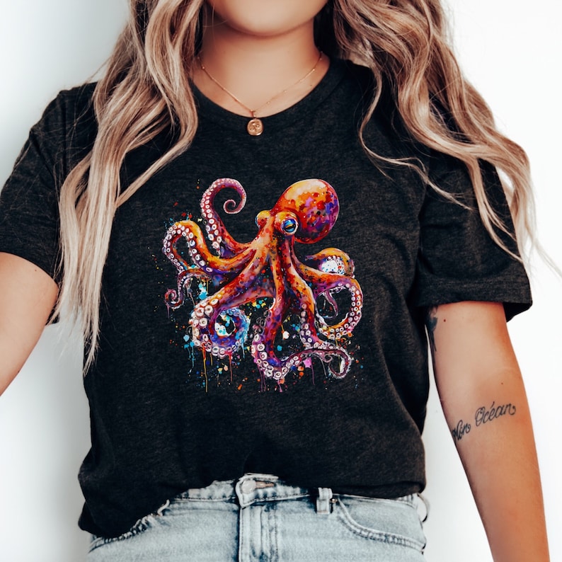 A black t-shirt with a vibrant, detailed graphic of an octopus. The octopus is rendered in a watercolor or paint splatter style with a mix of bright colors, including orange, purple, blue, and yellow. The design features splatters and drips of paint around the tentacles. The shirt is worn by a person with long, blonde hair, a gold necklace, and a small tattoo on their left arm.