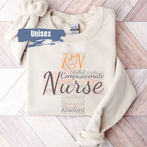 May include: A beige unisex sweatshirt with a word cloud design featuring the words "RN" and "Nurse" in a peach color. The word cloud also includes words like "skilled", "compassionate", "reliable", "caring", "empathetic", "dedicated", and "resilient".