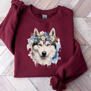 Op de afbeelding: Bordeauxkleurige sweatshirt met een aquarel illustratie van een husky hond met een bloemenkrans. De hond heeft wit en grijs haar, met blauwe en witte bloemen in de krans. Het ontwerp is geplaatst tegen een blauwe aquarel splash.