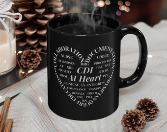 May include: A black coffee mug with a white heart-shaped design filled with medical terms, including "CDI At Heart." Steam rises from the mug, suggesting a hot beverage. The mug is on a light-colored surface with decorative elements.