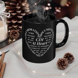 May include: A black coffee mug with a white heart-shaped design filled with medical terms, including "CDI At Heart." Steam rises from the mug, suggesting a hot beverage. The mug is on a light-colored surface with decorative elements.