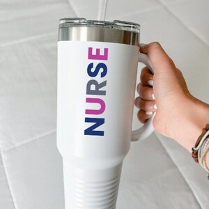 May include: White insulated tumbler with a silver rim and handle. The word "NURSE" is printed vertically in pink and navy blue. The tumbler is designed to keep beverages hot or cold.