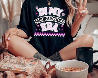 May include: Black t-shirt with the text "IN MY MEDICAL CODER ERA" in pink and white. The design includes floral and geometric accents. The shirt is worn by a person sitting cross-legged.