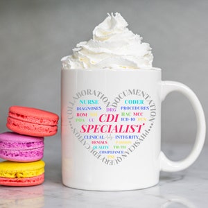 May include: A white ceramic mug with a heart-shaped design filled with medical terms, including "CDI Specialist." The mug is topped with whipped cream. Three colorful macarons are stacked to the left of the mug.