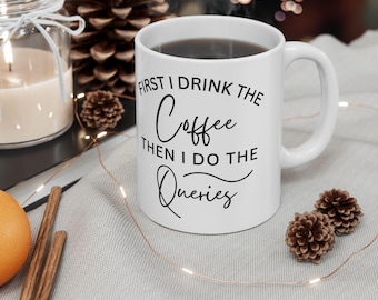 May include: White ceramic coffee mug filled with dark liquid, featuring the text "FIRST I DRINK THE Coffee THEN I DO THE Queries" in black script. The mug has a curved handle and is surrounded by decorative elements.