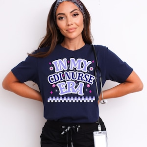 May include: Navy blue t-shirt with the text "IN MY CDI NURSE ERA" in a purple and white design. The shirt has a decorative border and is likely made of a soft, comfortable material. A stethoscope hangs around the neck.