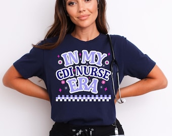May include: Navy blue t-shirt with the text "IN MY CDI NURSE ERA" in a purple and white design. The shirt has a decorative border and is likely made of a soft, comfortable material. A stethoscope hangs around the neck.