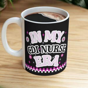 May include: White ceramic coffee mug with a black background featuring the text "IN MY CDI NURSE ERA" in pink and white. The mug is filled with dark coffee and has a white handle.