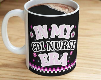 May include: White ceramic coffee mug with a black background featuring the text "IN MY CDI NURSE ERA" in pink and white. The mug is filled with dark coffee and has a white handle.
