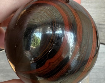 May include: A polished, red and black, tiger's eye sphere on a clear stand. The sphere has a swirling pattern of red and black.