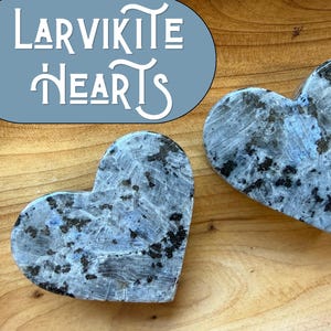 Könnte beinhalten: Zwei herzförmige Steine aus Larvikit mit einer polierten Oberfläche und einem gesprenkelten Muster aus grauen, blauen und schwarzen mineralischen Einschlüssen. Der Text "LARVIKITE HEARTS" steht in einer blauen ovalen Grafik oben links.