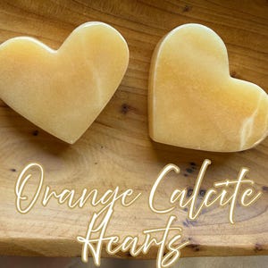 Könnte beinhalten: Zwei glatte, polierte herzförmige Steine aus orangem Calcit. Die Steine haben einen durchscheinenden, warmen gelb-orangen Farbton mit subtiler natürlicher Maserung. Der Text "Orange Calcite Hearts" ist in weißer Schreibschrift darübergelegt.