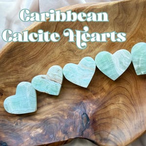 Könnte beinhalten: Fünf herzförmige Steine aus karibischem Calcit mit hellgrünen und weißen Streifenmustern. Die Steine sind in einer diagonalen Linie in einer Schale aus poliertem Holz angeordnet. Der Text "Caribbean Calcite Hearts" ist in Weiß darübergelegt.