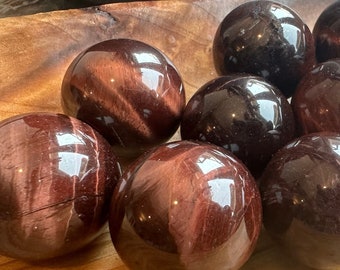 May include: A collection of 12 red tiger's eye spheres in a wooden bowl. The spheres are polished and have a reddish-brown color with a golden sheen.