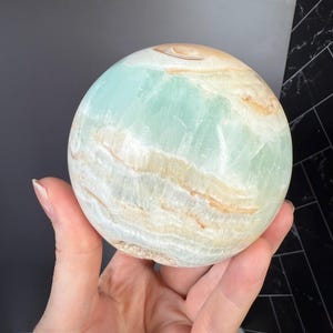 May include: A polished sphere of Caribbean calcite, showcasing a blend of pale blue, cream, and tan hues. The gemstone's surface displays natural banding and a smooth, rounded form. This decorative sphere is a unique home decor item.