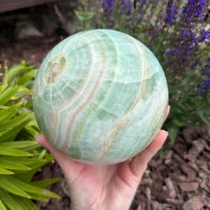 May include: A polished spherical stone featuring a smooth surface with swirling patterns of light mint green, pale cream, and soft white bands. The orb displays natural mineral inclusions and translucent layers throughout its rounded form.