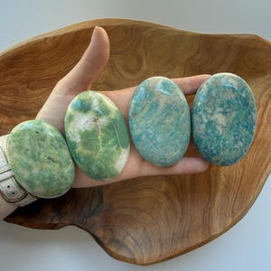 May include: Four smooth, oval-shaped polished stones in varying shades of green and teal. The stones exhibit natural mineral patterns with white inclusions and glossy surfaces, arranged in a row to display their distinct color variations.