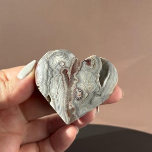 May include: Heart-shaped stone carving with a layered pattern of gray, white, and brown. The polished surface reveals intricate bands and swirls, creating a natural, organic design. The stone is held in a hand.