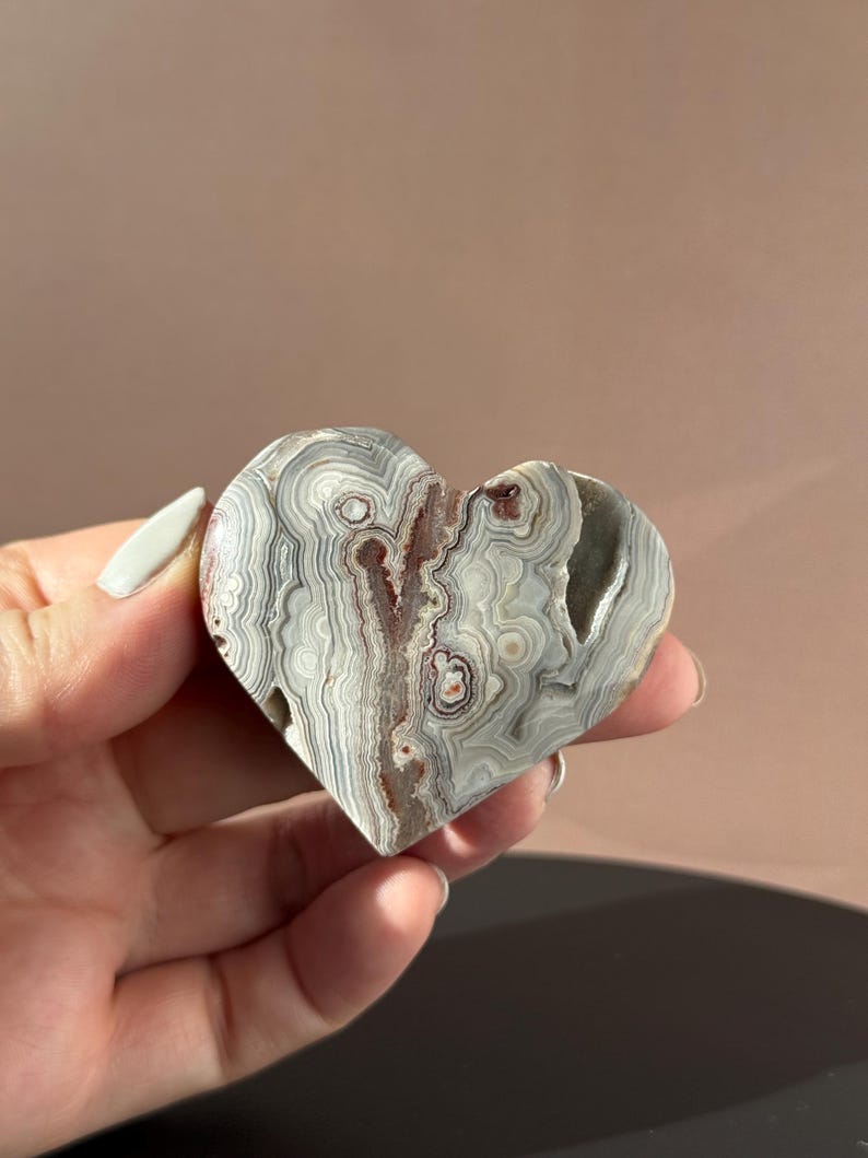 May include: Heart-shaped stone carving with a layered pattern of gray, white, and brown. The polished surface reveals intricate bands and swirls, creating a natural, organic design. The stone is held in a hand.