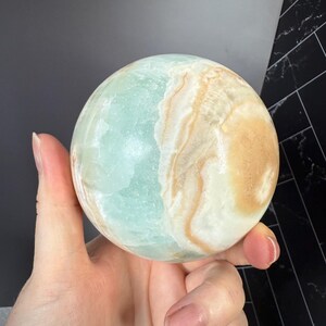 K&ouml;nnte beinhalten: Eine polierte Kugel aus karibischem Calcit, die eine Mischung aus hellblauen, cremefarbenen und beigefarbenen Farbt&ouml;nen aufweist. Die Kugel wird in einer Hand gehalten, wodurch die nat&uuml;rlichen Muster und Texturen des Steins hervorgehoben werden. Die Kugel ist ein dekorativer Gegenstand.