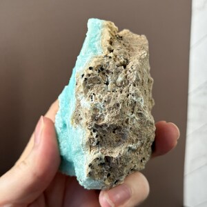 Puede incluir: Una mano sostiene un esp&eacute;cimen mineral natural. La roca presenta un contraste llamativo entre un azul turquesa claro y una textura rugosa, porosa, marr&oacute;n-beige. La superficie del mineral es irregular, mostrando su formaci&oacute;n natural.