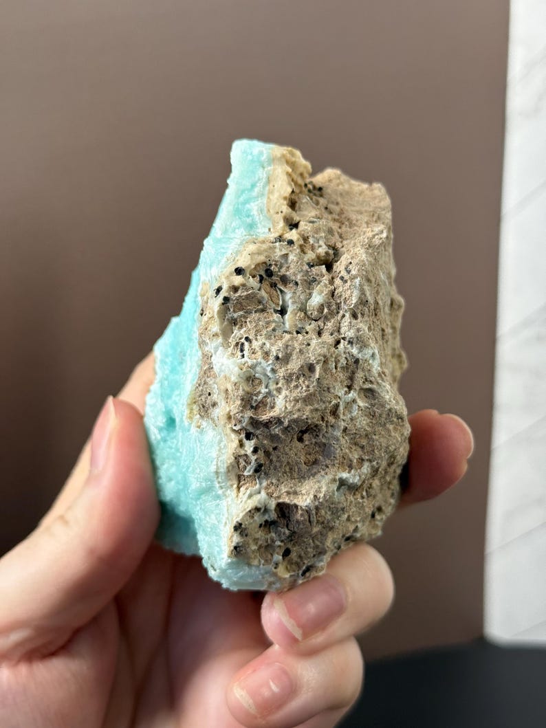 Puede incluir: Una mano sostiene un esp&eacute;cimen mineral natural. La roca presenta un contraste llamativo entre un azul turquesa claro y una textura rugosa, porosa, marr&oacute;n-beige. La superficie del mineral es irregular, mostrando su formaci&oacute;n natural.