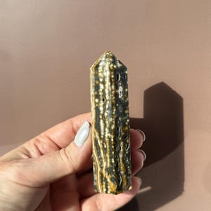 Puede incluir: Un obelisco de piedra preciosa pulida con una parte superior puntiaguda. La piedra presenta un patr&oacute;n de manchas y rayas negras, blancas y doradas. El obelisco se sostiene en una mano sobre un fondo neutro.