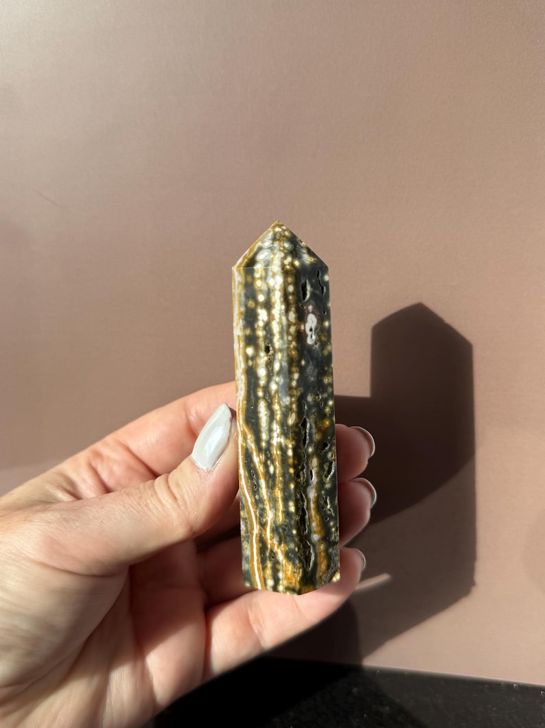 Puede incluir: Un obelisco de piedra preciosa pulida con una parte superior puntiaguda. La piedra presenta un patr&oacute;n de manchas y rayas negras, blancas y doradas. El obelisco se sostiene en una mano sobre un fondo neutro.