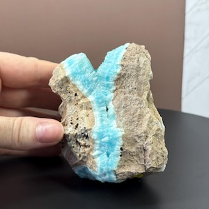 May include: A rough, natural stone with a striking vein of bright turquoise blue running through it. The stone's exterior is a mix of beige and brown, with a textured surface. The blue vein adds a vibrant contrast to the earthy tones of the rock.