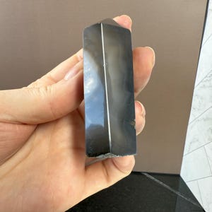May include: A polished, rectangular, dark gray crystal tower with white and gray veining. The crystal is held in a hand, showcasing its smooth, reflective surface. The tower has a flat top and a faceted design.