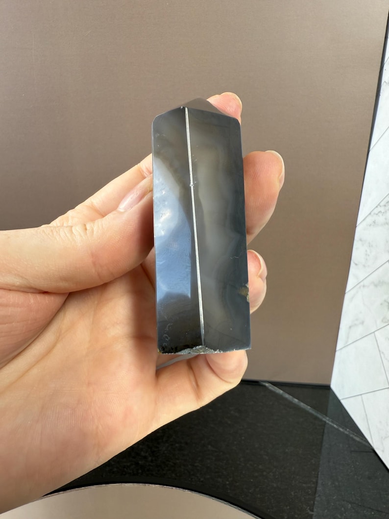 May include: A polished, rectangular, dark gray crystal tower with white and gray veining. The crystal is held in a hand, showcasing its smooth, reflective surface. The tower has a flat top and a faceted design.