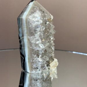 May include: A clear quartz crystal point with a banded agate base. The crystal has a natural, rough texture and a polished, reflective surface. The agate features bands of white, gray, and black. The crystal is standing upright.