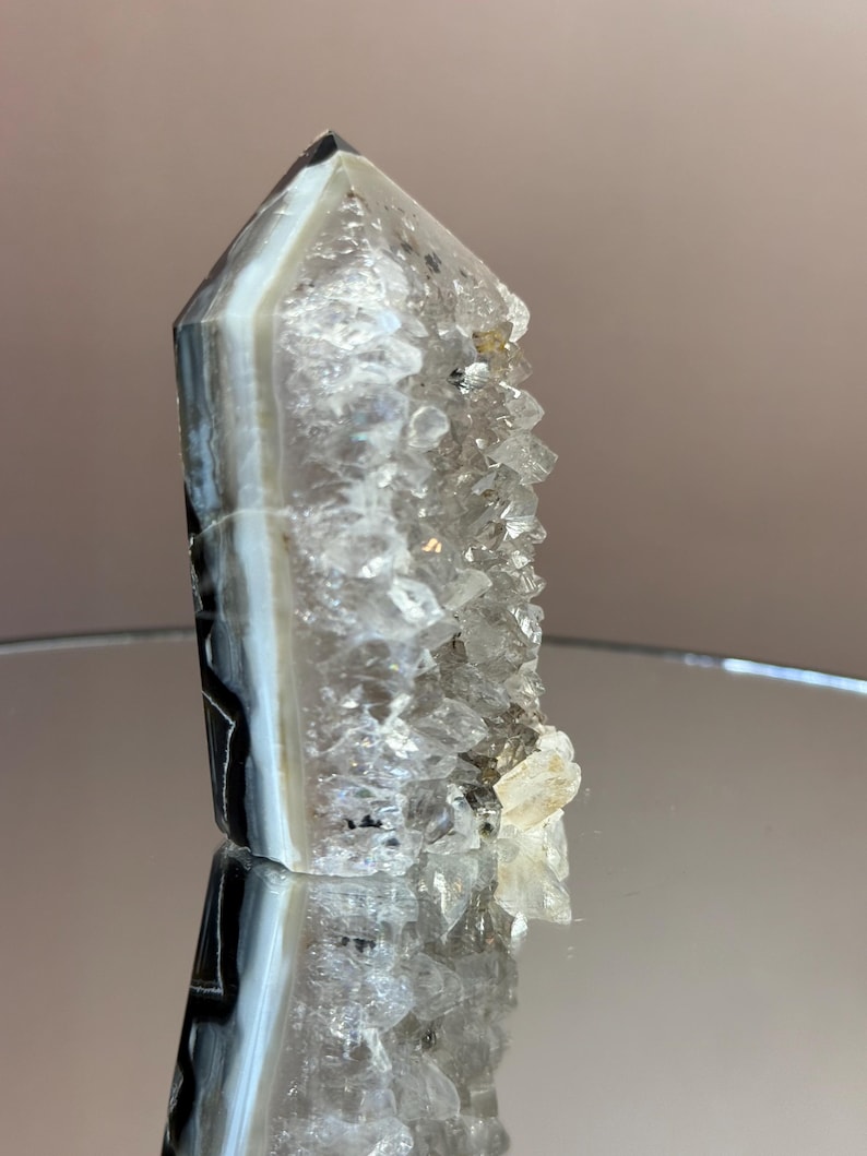May include: A clear quartz crystal point with a banded agate base. The crystal has a natural, rough texture and a polished, reflective surface. The agate features bands of white, gray, and black. The crystal is standing upright.