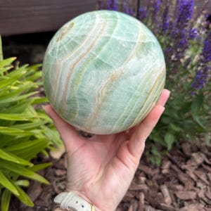 May include: A polished spherical stone featuring bands of pale mint green and soft cream white. The surface displays natural translucent layers and swirling mineral patterns throughout the round form, creating a smooth, glossy texture across the object.
