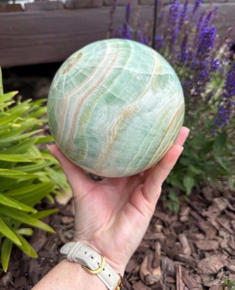 May include: A polished spherical stone featuring bands of pale mint green and soft cream white. The surface displays natural translucent layers and swirling mineral patterns throughout the round form, creating a smooth, glossy texture across the object.
