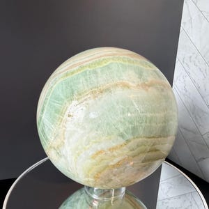 May include: A polished spherical stone sculpture featuring bands of pale green, cream, and soft orange hues. The surface displays natural mineral veining and translucent layers throughout the orb, creating a smooth, reflective finish on the rounded form.
