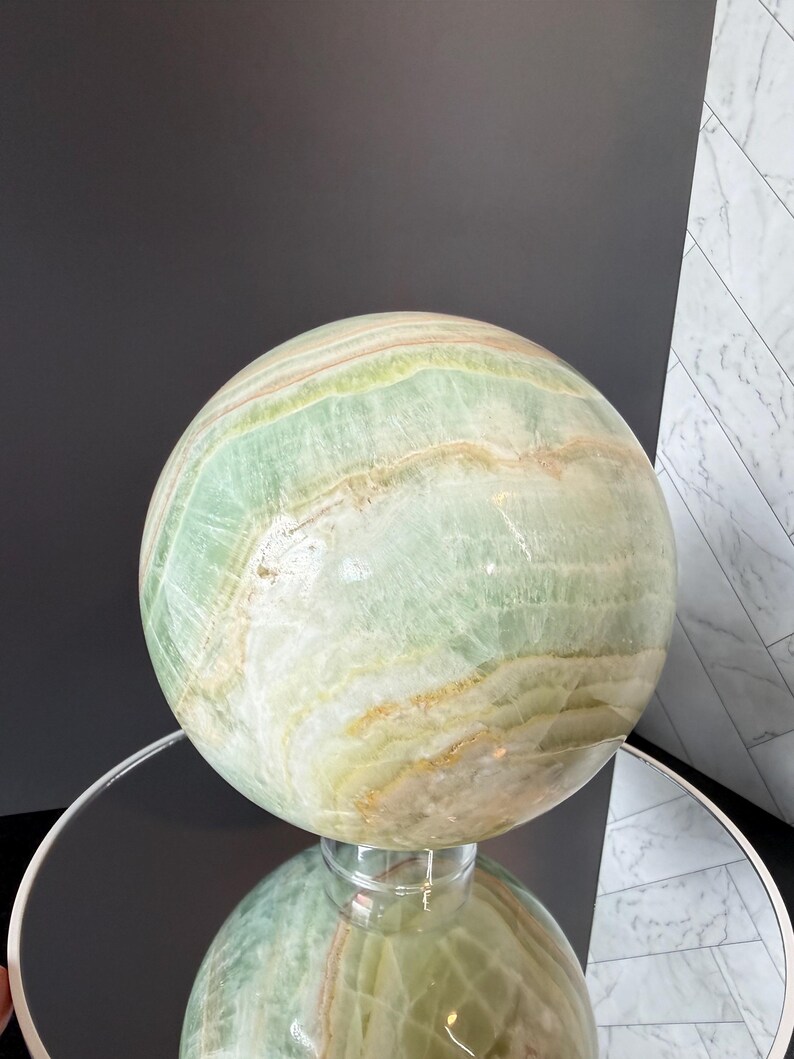 May include: A polished spherical stone sculpture featuring bands of pale green, cream, and soft orange hues. The surface displays natural mineral veining and translucent layers throughout the orb, creating a smooth, reflective finish on the rounded form.