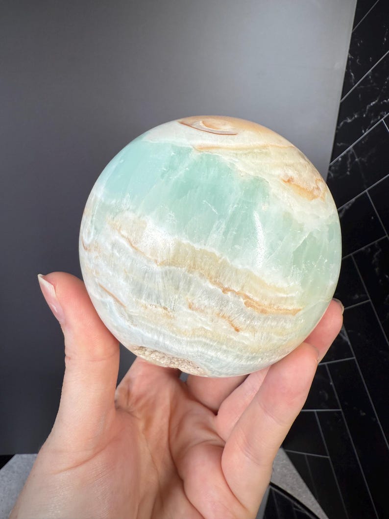 May include: A polished sphere of Caribbean calcite, showcasing a blend of pale blue, cream, and tan hues. The gemstone's surface displays natural banding and a smooth, rounded form. This decorative sphere is a unique home decor item.