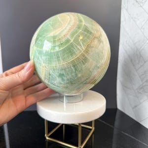May include: A polished green sphere with visible white and light brown banding patterns. The sphere rests on a clear circular stand, which is supported by a white circular platform and a gold-toned metal frame with four thin legs.