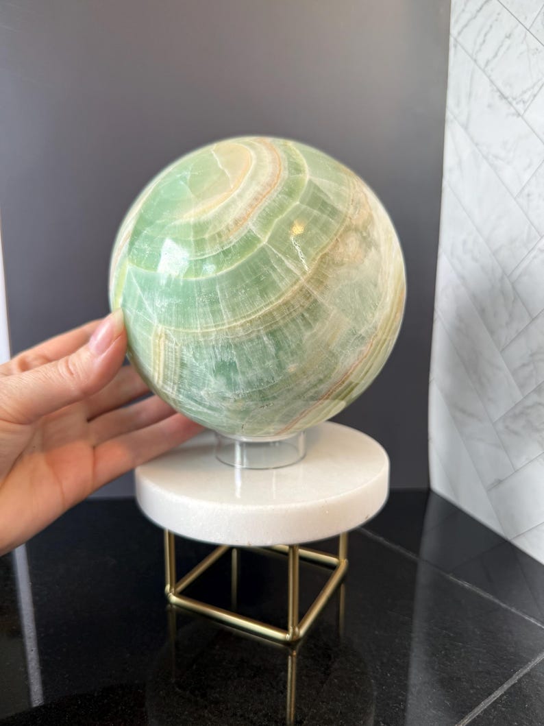 May include: A polished green sphere with visible white and light brown banding patterns. The sphere rests on a clear circular stand, which is supported by a white circular platform and a gold-toned metal frame with four thin legs.